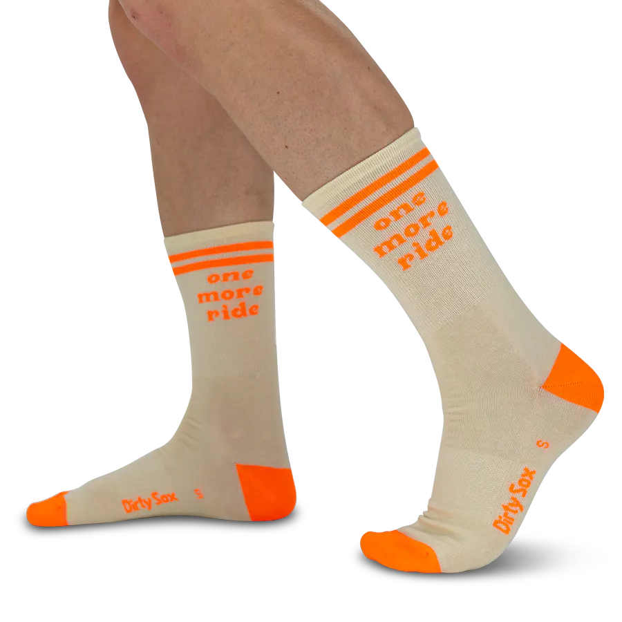Jambes en chaussettes de sport beiges avec détails orange vif : deux bandes horizontales à la bordure, puis l'inscription « one more ride ». Pointe et talon orange intense, contrastant fortement.