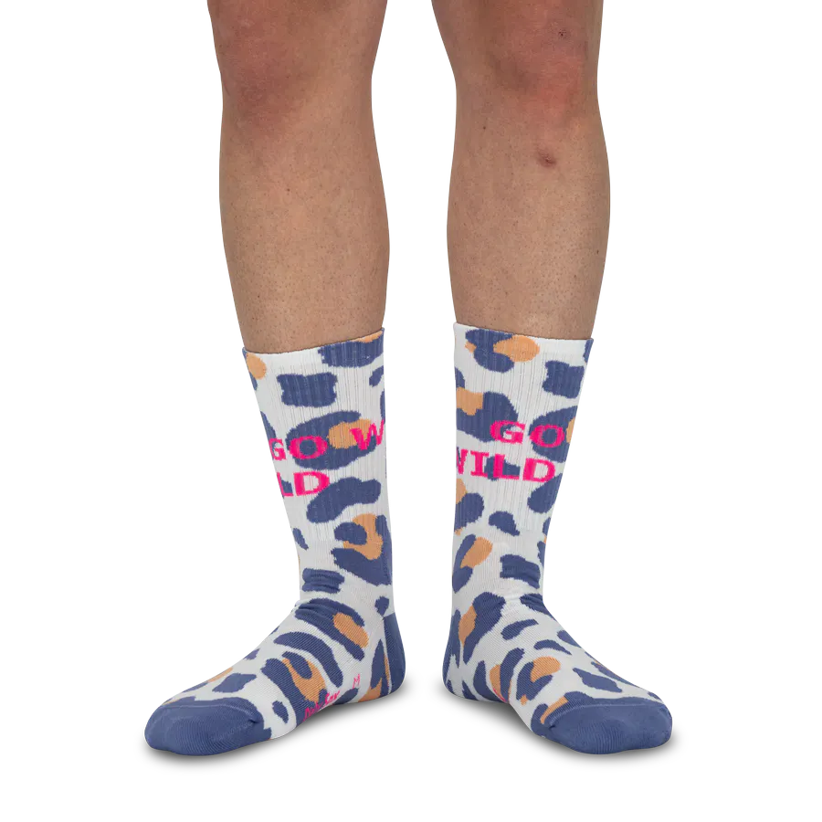 A person is wearing white socks featuring a vibrant blue and orange leopard print and the pink lettering ‘GO WILD’. The socks cover the ankles and reach up to the calves. The focus is on the feet and lower legs against a white background.