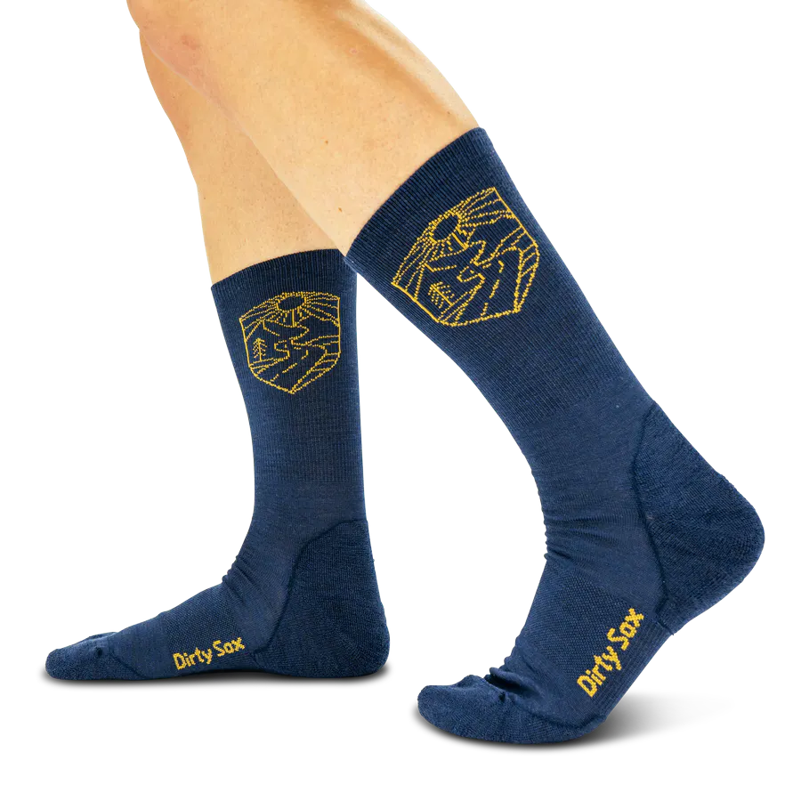 Zwei Beine tragen dunkelblaue Socken mit gelbem Emblem: stilisierte Landschaft mit Bergen, Bäumen und aufgehender Sonne am Horizont. Am Fuß steht „Dirty Sox“ in Gelb. Sportliche Passform, geeignet für Outdoor-Aktivitäten.