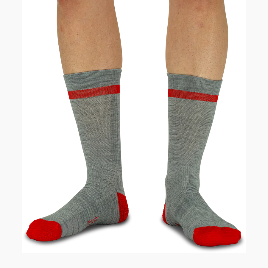 Ein Paar Waden trägt hohe, hellgraue Alpine Heritage Trekkingsocken. Ein roter Streifen ziert den Bund, Zehen und Fersen sind leuchtend rot. Das robuste Merinowoll-Strickmuster reguliert Temperatur und Feuchtigkeit auf langen Touren.
