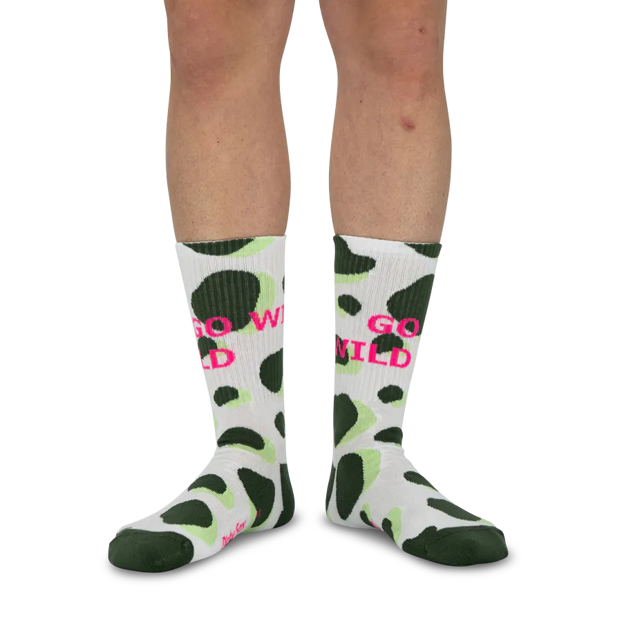 Une personne porte des chaussettes mi-mollet blanches à taches irrégulières vert foncé et vert clair, motif animalier (girafe). Devant, « GO WILD » en rose vif. Pieds vers l'avant, fond blanc.