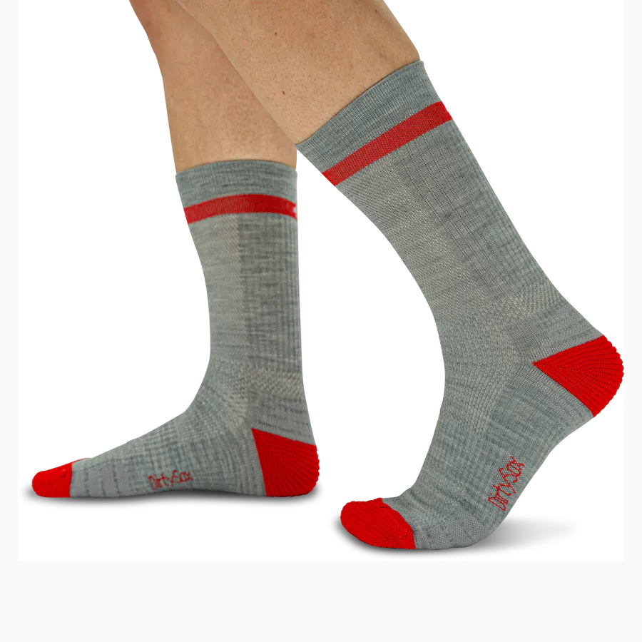 Graue Trekkingsocken aus Merinowolle werden von einer Person getragen. Sie reichen bis zur Wadenmitte und haben einen auffälligen roten Streifen am Schaft sowie leuchtend rote Fersen- und Zehenbereiche. DirtySox-Logo seitlich.