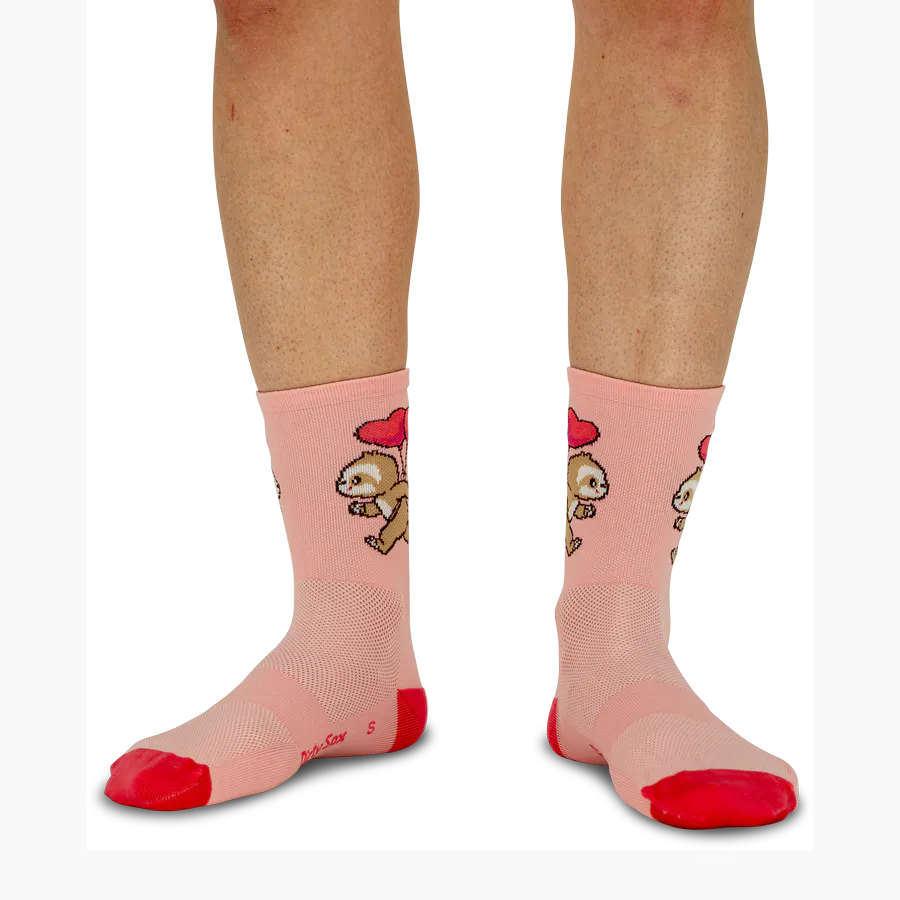 Zwei Beine auf weißem Hintergrund tragen pinke Running-Socken mit wiederholtem Faultiermuster, jedes hält einen roten Herzballon. Spitzen und Fersen sind leuchtend rot und betonen das sportlich-verspielte Design.