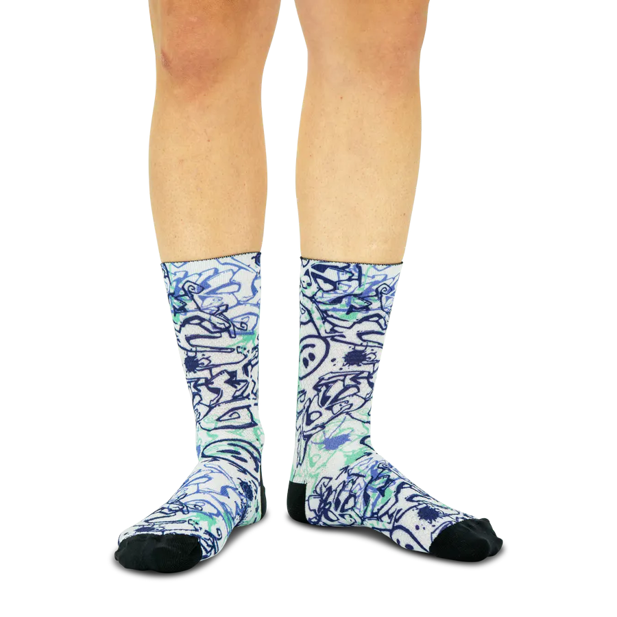 Zwei Beine tragen weiße Socken mit energiegeladenem Graffiti-Muster in Blautönen und frischem Mintgrün. Chaotische Formen und Spritzer heben sich ab, während Zehen und Fersen schwarz sind.