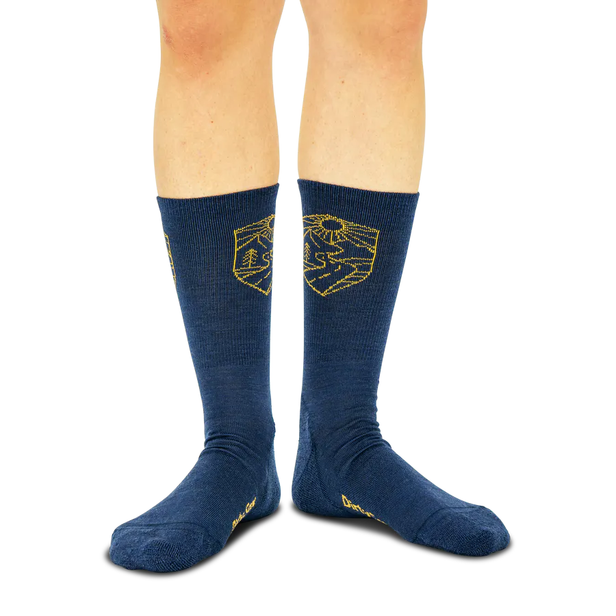 Zwei Beine in marineblauen Socken mit goldenen Emblemen. Links stilisierte Berge und Bäume. Rechts eine strahlende Sonne über dem Horizont mit gewundenem Flusslauf. Die ozeanblaue Farbe passt zum Outdoor-Design.
