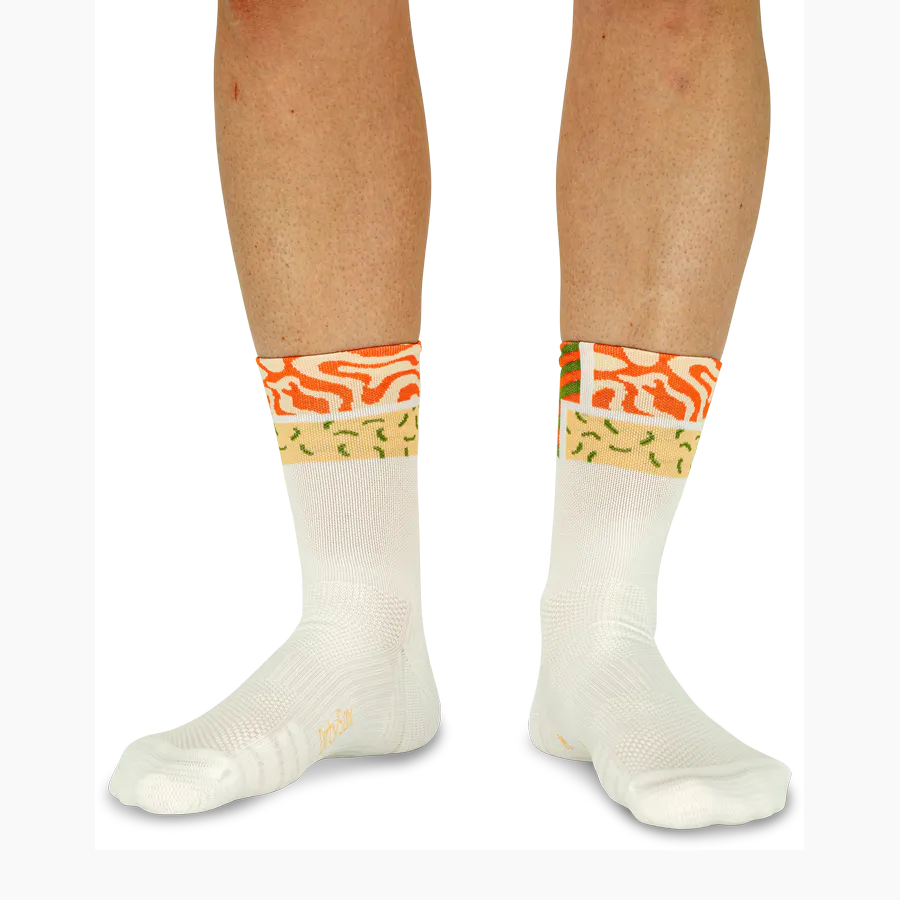 Ein Paar Beine trägt weiße Sportsocken. Der obere Bund zeigt ein lebhaftes `Tangerine Wave`-Design mit Wellen in Orangerot und Cremeweiß. Darunter ein hellgelber Streifen mit kleinen grünen Tupfen. Rest schlicht weiß bis zur Wadenmitte.