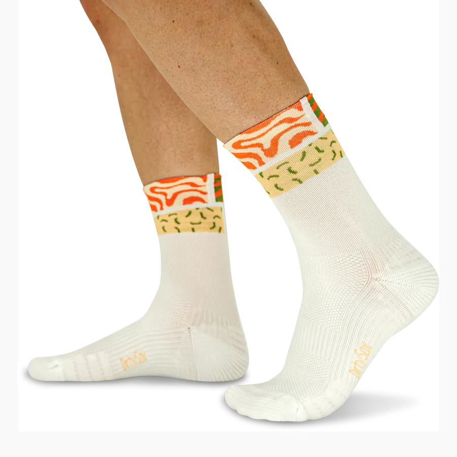 Zwei Beine in cremefarbenen Socken mit auffälligen Bündchenmustern: orangefarbene Wellenstruktur und hellgrüne Sprinkles auf gelbem Grund. Sportliche Socken mit feiner Textur, seitlich das „Dirty Sox“-Logo. Vor weißem Hintergrund.