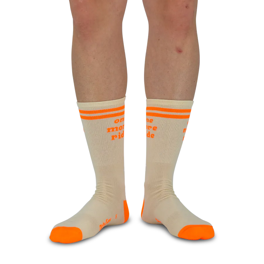 Une paire de jambes porte des chaussettes de sport crème avec détails orange vif. Chaque chaussette a deux bandes orange en haut et l’inscription « one more ride » en orange. Orteils et talons orange.