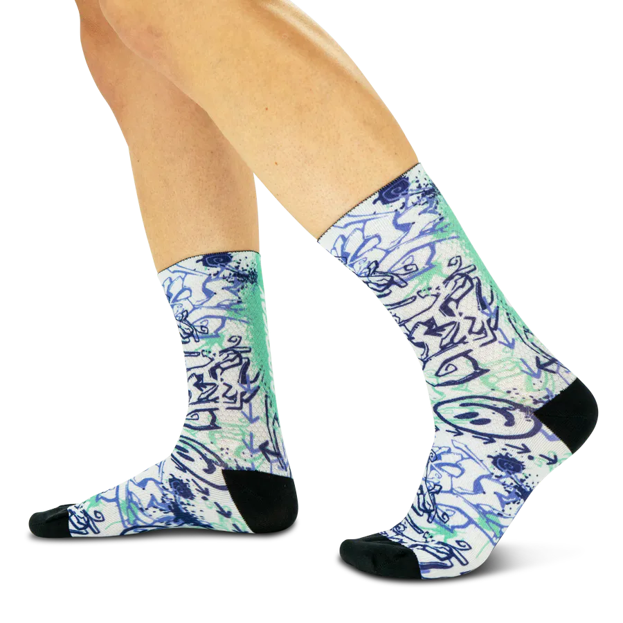 Ein Paar Beine trägt weiße Socken mit einem lebhaften Graffiti-Muster in Blau- und Minttönen. Das abstrakte Design zeigt verschlungene Linien und Farbspritzer, die an urbane Kunst erinnern. Die Socken haben schwarze Fersen und Zehenbereiche.