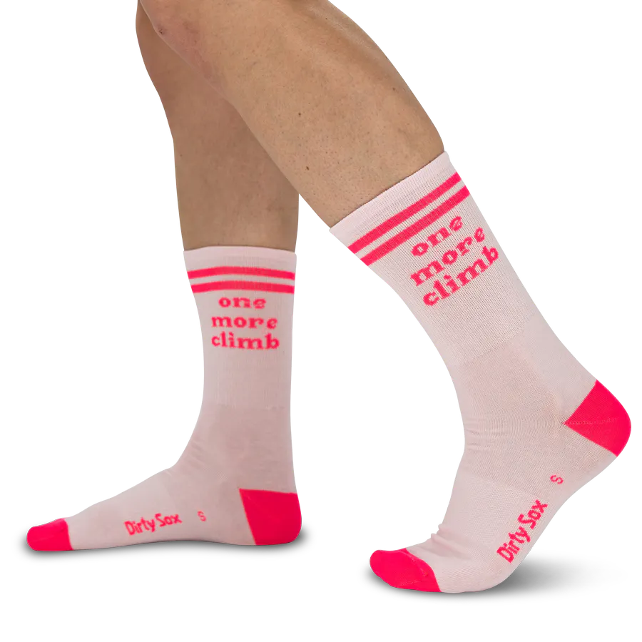 Zwei Beine tragen hellrosa Socken mit neonpinken Akzenten. Nahe des Bündchens sind drei horizontale Streifen und der Schriftzug 'one more climb' in leuchtendem Pink zu sehen. Zehen und Fersen sind ebenfalls neonpink, und an der Seite steht 'Dirty Sox'.